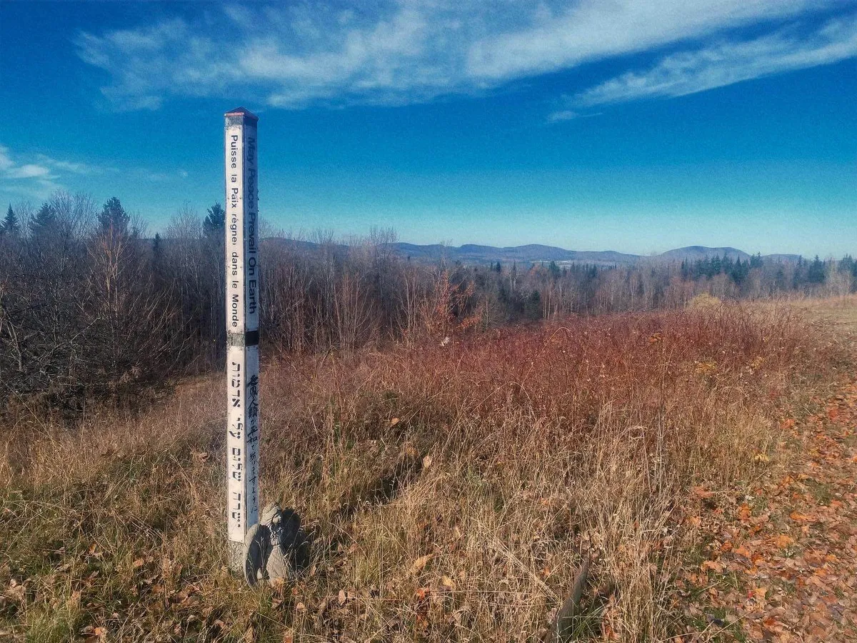 A wide, open field stretches out with a clearly visible trail marker post in the foreground, leading the eye toward distant blue mountains. Soft early morning or late afternoon light casts a serene glow, hinting at the possibilities for hiking, nature walks, and group adventures. The openness of the landscape is inviting for exploration and large gatherings.
