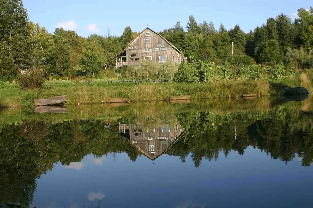 A stunning exterior view of a rustic house overlooking a calm pond, surrounded by lush greenery. The tranquil setting suggests a peaceful spot for fishing, picnicking, or gathering as a group to enjoy sunsets or early mornings together.