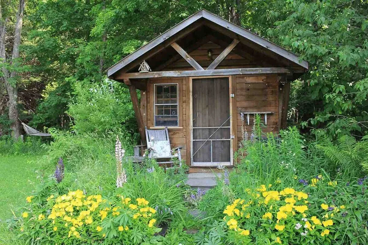 A wooden cabin surrounded by a vibrant flower garden, with a welcoming front porch bathed in natural light. The peaceful scene hints at morning activities such as enjoying coffee outdoors or relaxing while immersed in nature.
