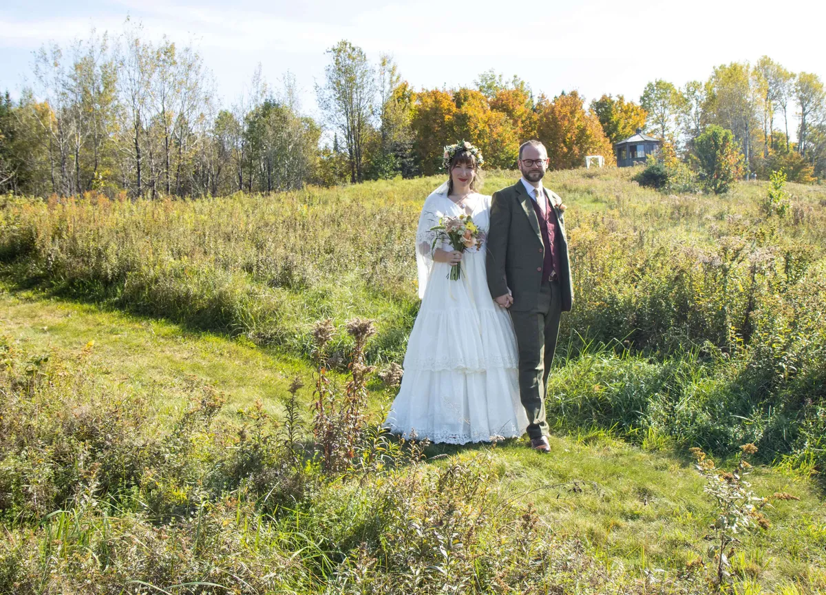 An exterior shot of a bride and groom embracing among wildflowers and autumn-colored trees. The couple stands in the foreground, radiating joy, with vibrant nature enveloping them. The mood is romantic and celebratory, emphasizing the creation of lasting memories in a picturesque setting.