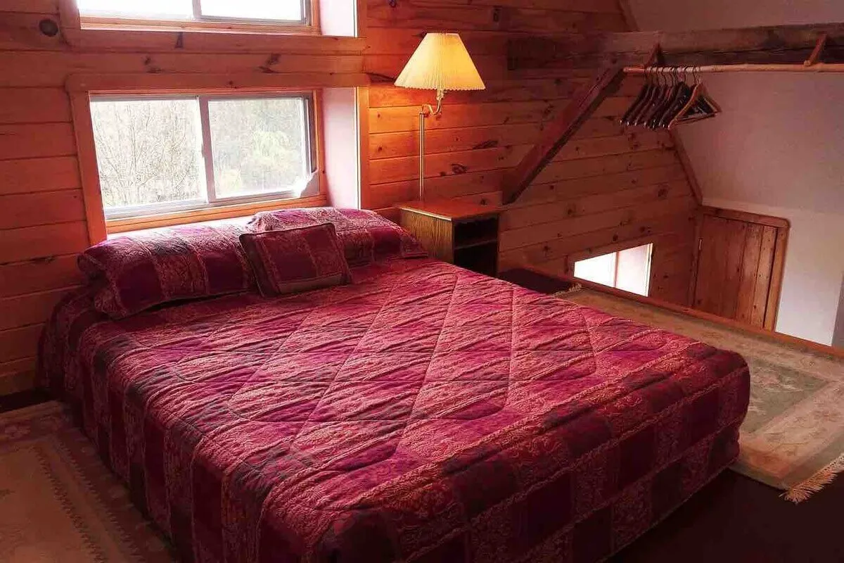 A cozy bedroom with wooden walls, a quilted bed, a warm lamp, and windows that let in soft natural light. The space feels peaceful and inviting, ideal for rest and relaxation after a day of activities.
