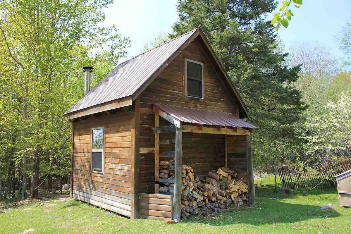 The image features a cozy wooden cabin with a stack of firewood outside, nestled in greenery. The welcoming porch hints at relaxed evenings, possibly spent around a fire pit, enhancing the sense of togetherness and outdoor relaxation.
