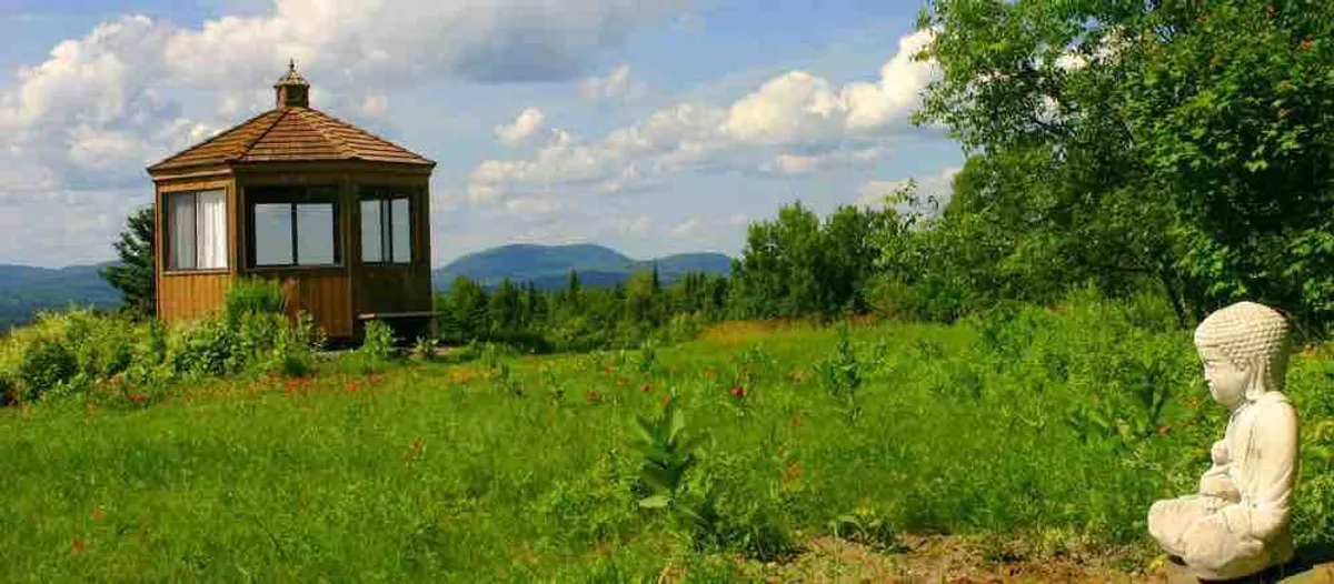 A peaceful outdoor gazebo featuring a Buddha statue, surrounded by lush greenery and a backdrop of gentle mountains. The space is designed for meditation and relaxation, evoking the ambiance of a purpose-built wellness retreat.