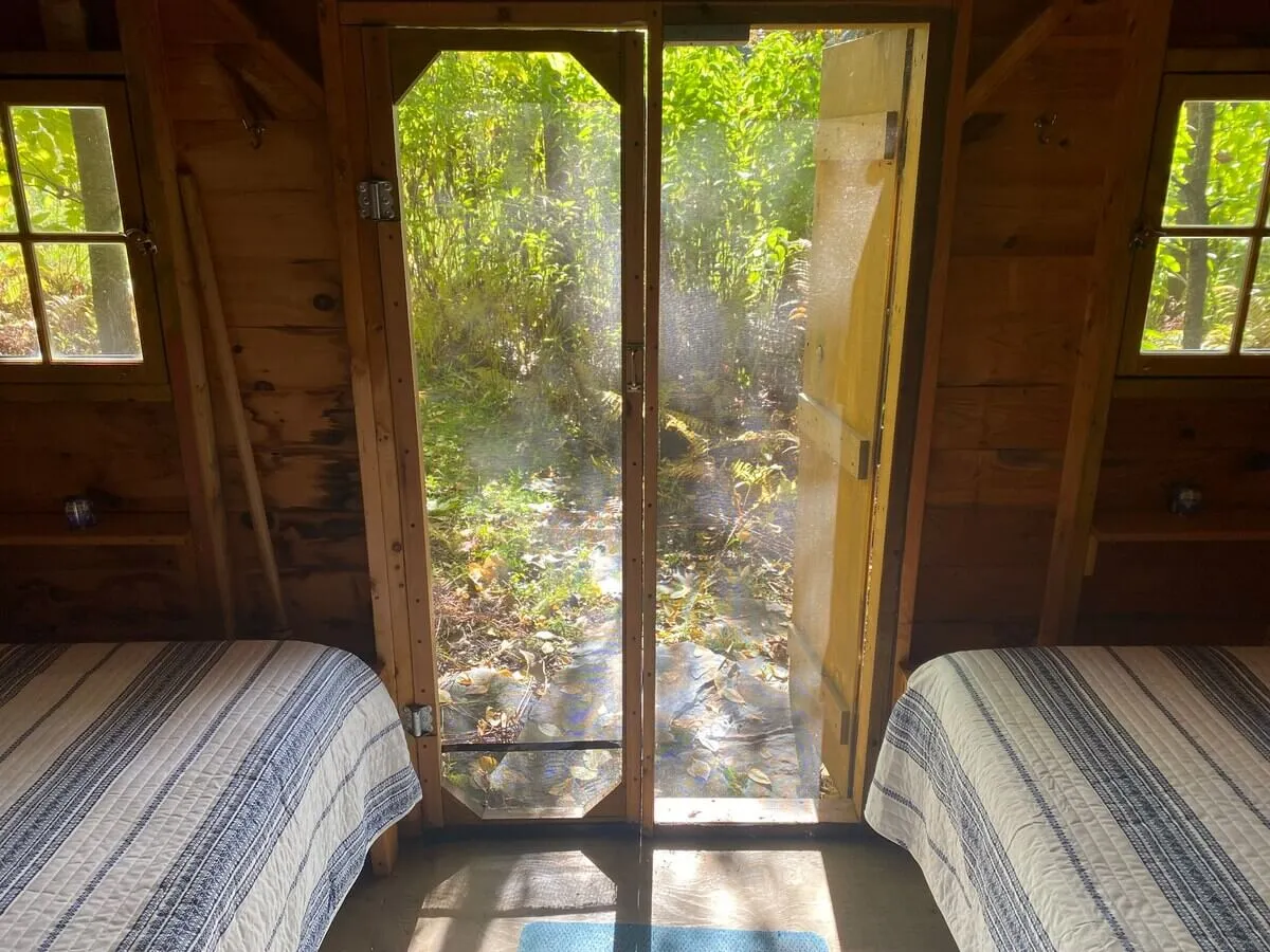 A peaceful room with a bed adorned in striped bedding, an open door letting in light and offering a view of the surrounding nature. The space feels both connected to the outdoors and secluded for restful privacy.