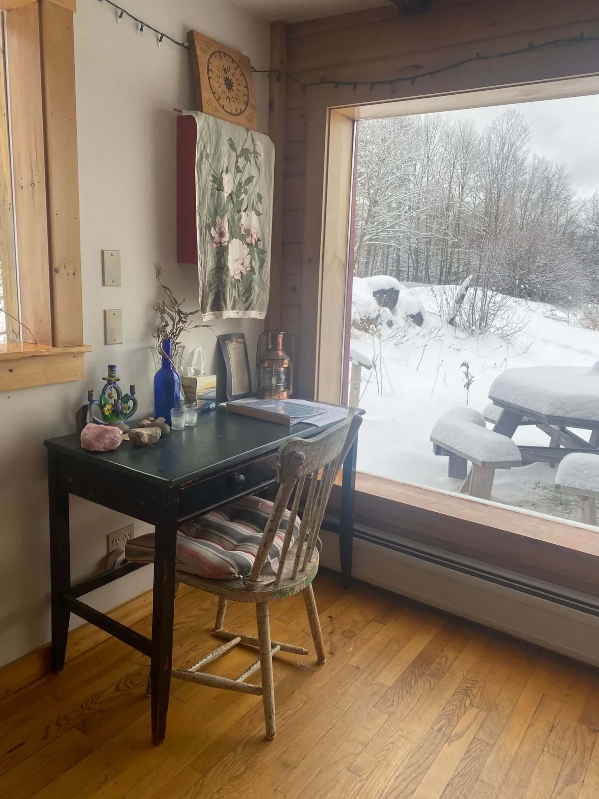 A tranquil interior featuring a wooden desk, large window, decorative wall hanging, and a chair. The soft natural light highlights the peaceful mood, making it an ideal spot for quiet reflection or solitary meditation.