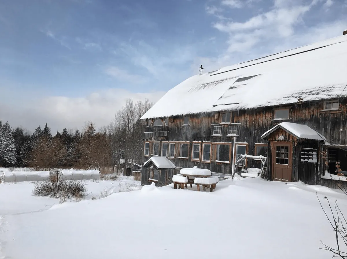 A snow-covered rustic barn stands amidst trees and a wintry landscape. Wooden tables and gathering areas hint at versatility for both indoor and outdoor events, even in colder months. The scene conveys a peaceful, rustic charm and shows the barn’s capacity to host cozy gatherings regardless of weather.