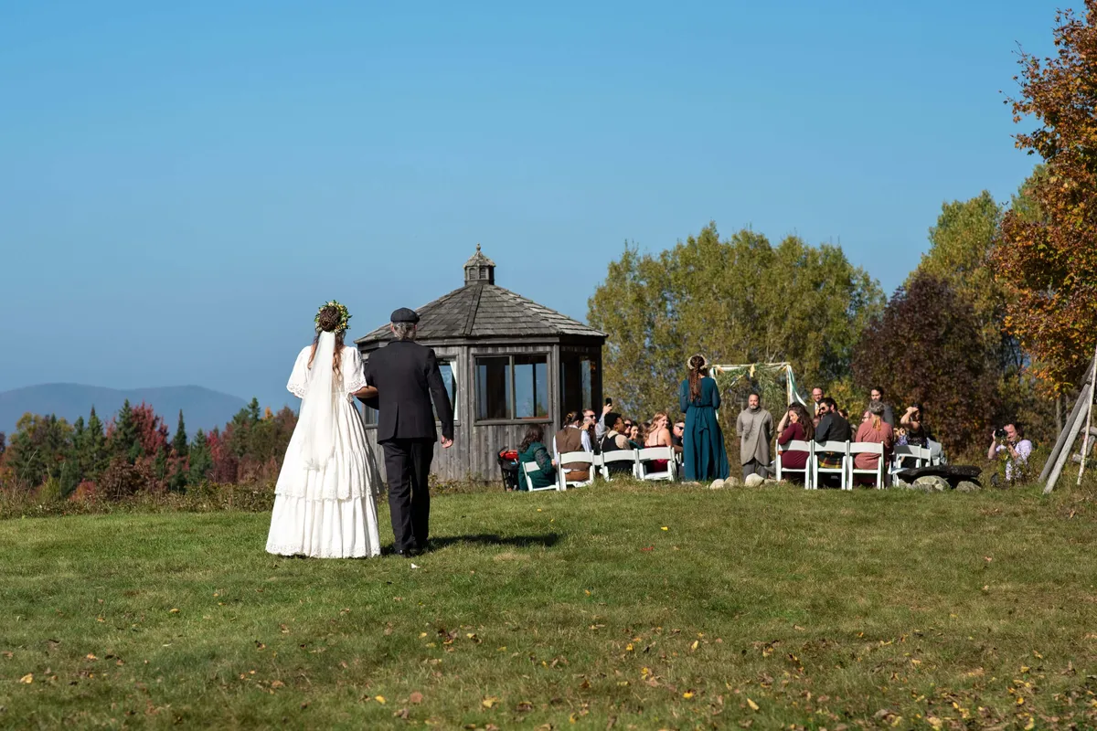 This image features an outdoor wedding ceremony with a bride and groom under a gazebo, surrounded by wedding guests. The picturesque setting is lush and green, framed by distant mountains, and the mood is joyful and celebratory. The scene highlights the emotional exchange of vows and the communal ambiance of a nature-inspired event.