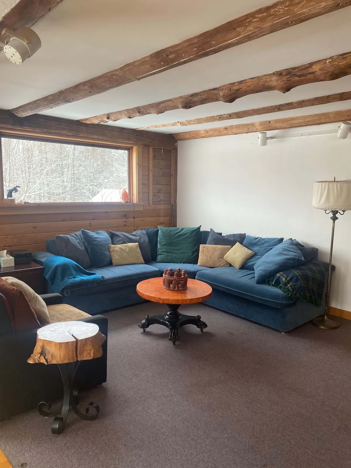 A cozy communal living room with a large sectional sofa, a central wooden coffee table, a standing lamp, and a panoramic window offering an inviting view of the outdoors. The arrangement encourages conversation, relaxation, and group activities such as reading or storytelling.