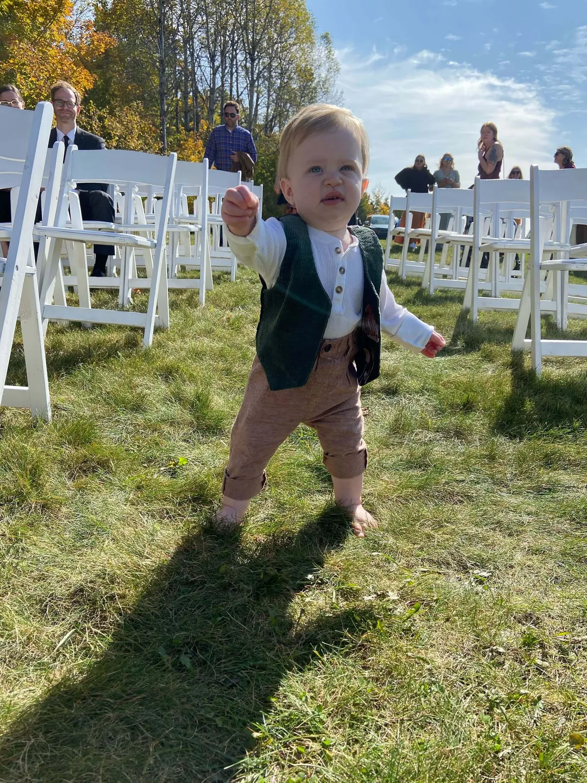 This exterior image captures a toddler taking first steps on a lawn surrounded by white chairs and autumn trees, with several people in the background. The playful, candid scene evokes family bonding and the joy of multi-generational gatherings in a natural outdoor setting.