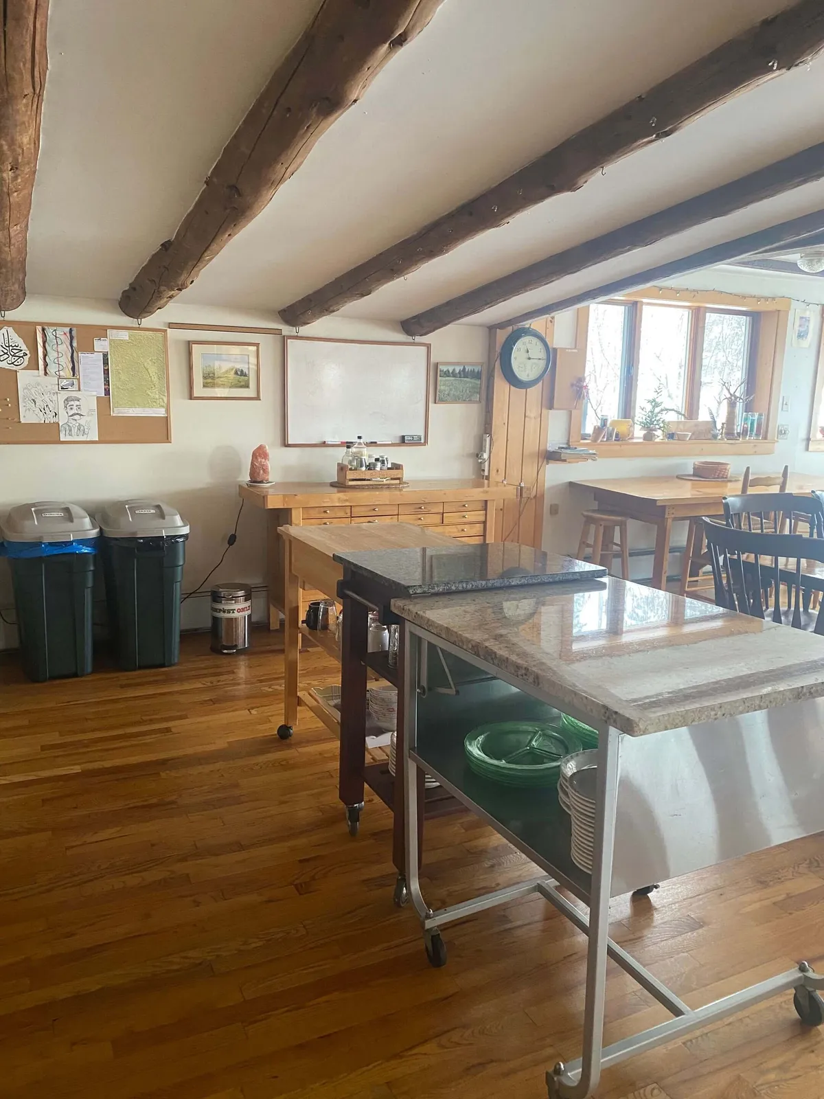 A spacious, light-filled kitchen featuring wooden beams, a central granite-topped island, ample counter space, a bulletin board for group planning, and large windows that bathe the space in natural light. The layout encourages communal cooking, meal prep, and group activities, fostering a warm, inviting atmosphere.