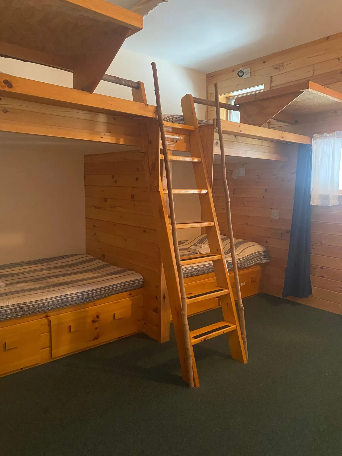 A playful, rustic bedroom featuring bunk beds with striped bedding, a ladder, and wooden walls. Natural light streams in, creating a warm and communal atmosphere perfect for groups or families.